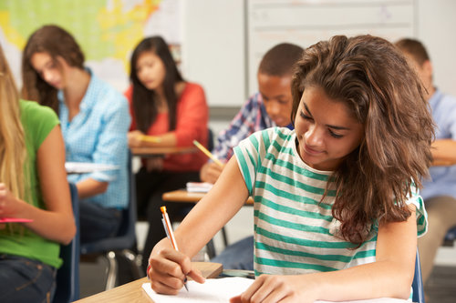 Female Pupil Studying At Desk In Classroom Sitting At Desk Writing In Textbook